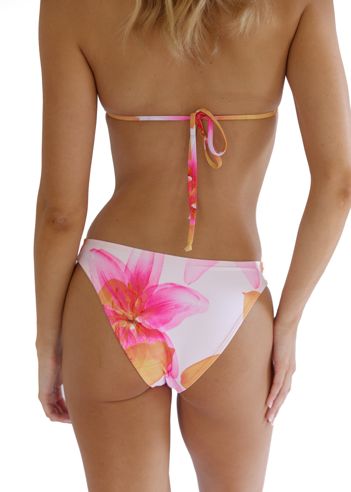 Back of a woman with blonde hair wearing a Lily Top back tie and Lily Midi Bottoms both in vibrant pink floral pattern, bottoms with mid-coverage, head to thighs visible, hands relaxed at sides on a white backdrop.