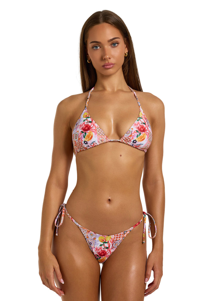 Woman with dark brown hair wearing a colorful La Mer Top in classic triangle halter style with adjustable neck and back ties and La Mer Bottoms both with a vibrant patchwork of floral and geometric patterns, including fruits, flowers, tiles, on a white backdrop, head to thighs visible, hands relaxed at sides on a white backdrop.