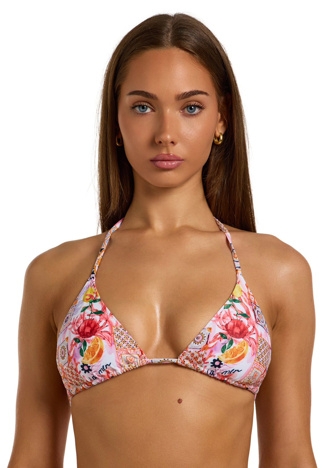 Woman with dark brown hair wearing a colorful La Mer Top in classic triangle halter style with adjustable neck and back ties with a vibrant patchwork of floral and geometric patterns, including fruits, flowers, tiles, and "la mer" wordings on pink background, with removable padding, on a white backdrop, head to torso shot.
