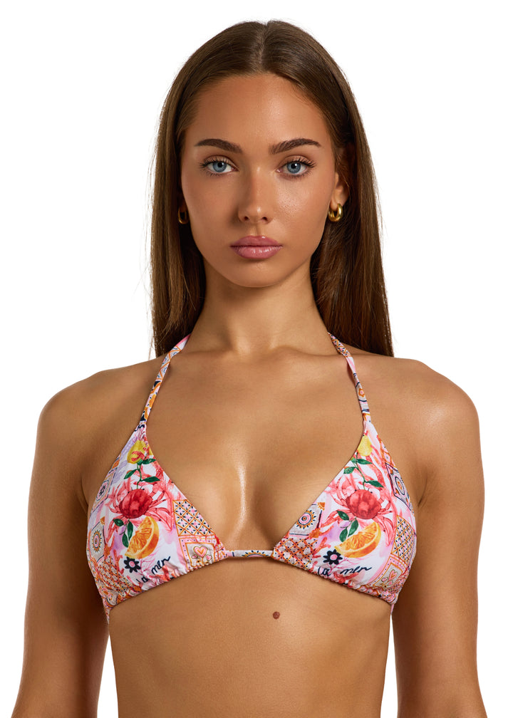 Woman with dark brown hair wearing a colorful La Mer Top in classic triangle halter style with adjustable neck and back ties with a vibrant patchwork of floral and geometric patterns, including fruits, flowers, tiles, and "la mer" wordings on pink background, with removable padding, on a white backdrop, head to torso shot.
