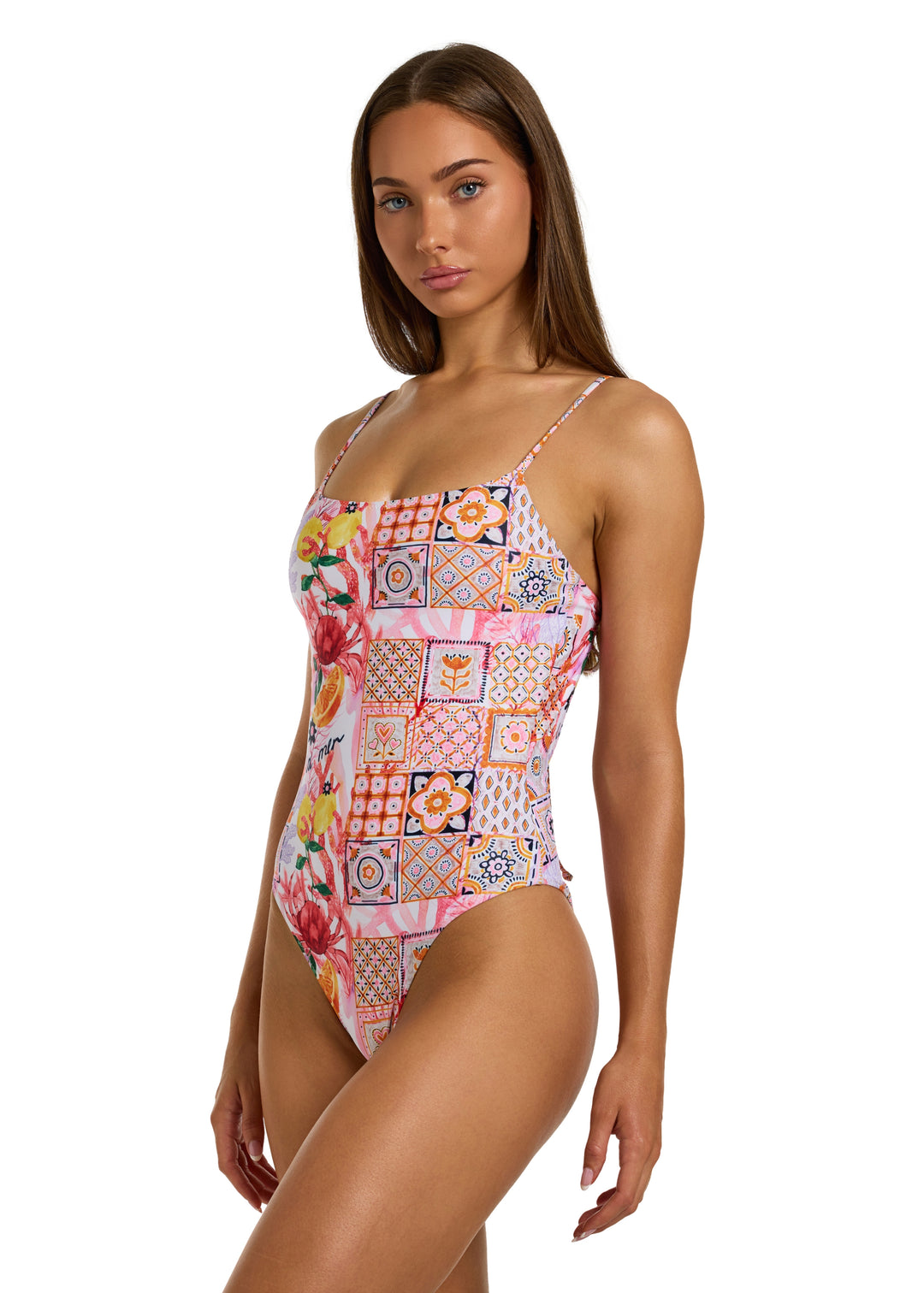 Front view of a woman with dark brown hair a bit facing left wearing the colorful La Mer One Piece swimsuit with thin straps, featuring a vibrant patchwork of floral and geometric patterns, including fruits, flowers, tiles, and the word "la mer" on pink background, non-padded style on a white backdrop, against a plain white backdrop, shown head to just above the knees with the left thigh slightly protruding to the left, on a white backdrop.