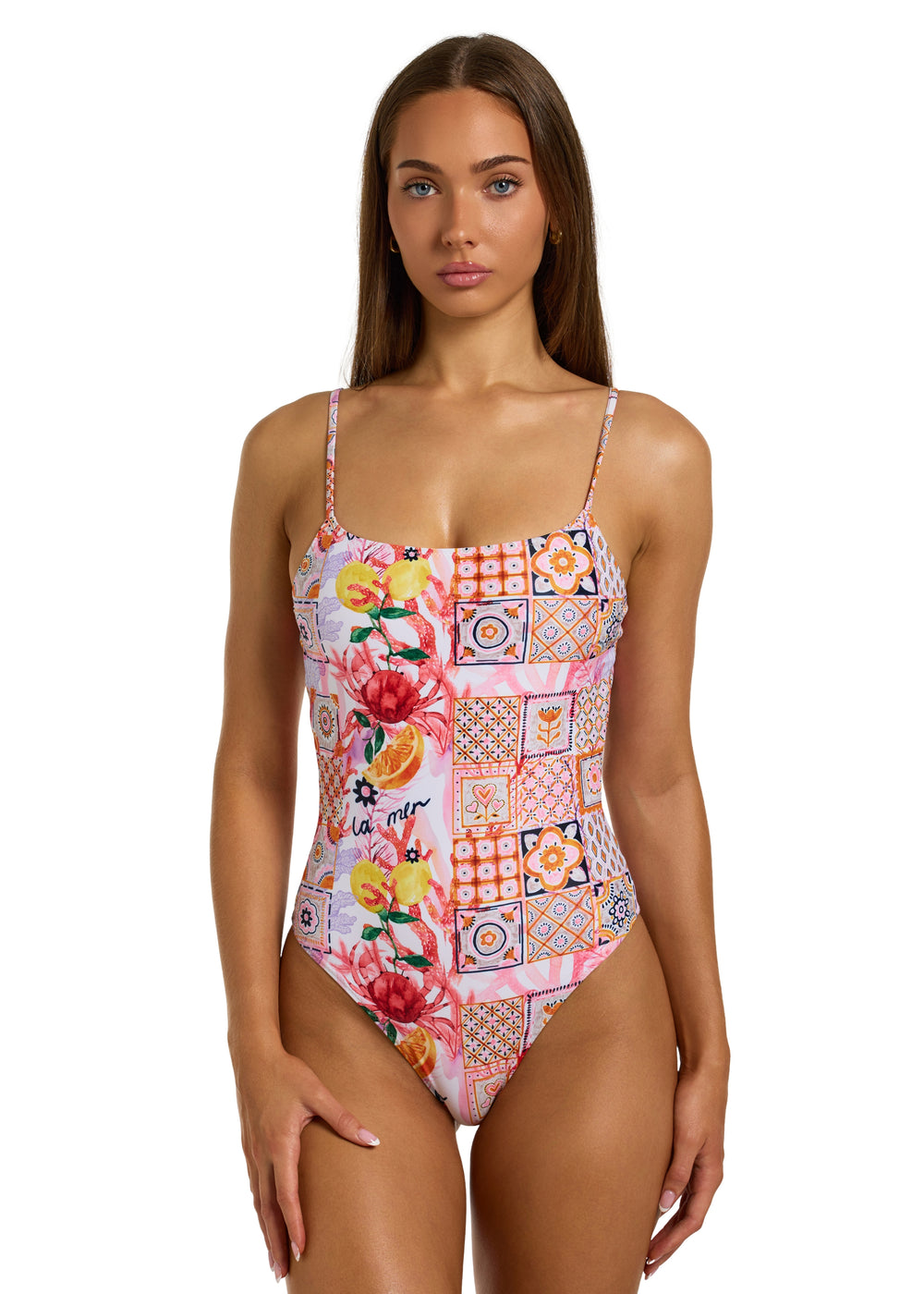 Front view of a woman with dark brown hair wearing the colorful La Mer One Piece swimsuit with thin straps, featuring a vibrant patchwork of floral and geometric patterns, including fruits, flowers, tiles, and the word "la mer" on pink background, non-padded style on a white backdrop, against a plain white backdrop, head to just above the knees visible, right hand resting on right thigh.