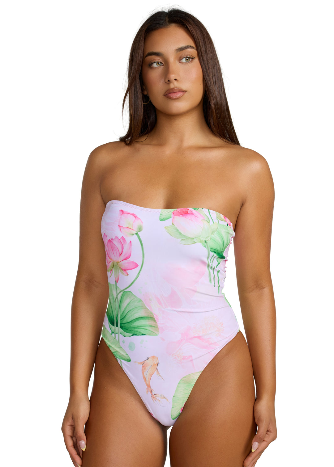 Front view of a woman with dark hair, eyes glancing left, wearing Koïa One Piece swimsuit bandeau style with light orange koi fish and soft pink and green lotus flower prints, visible from head to above the knees, hands resting on sides, against a white background.