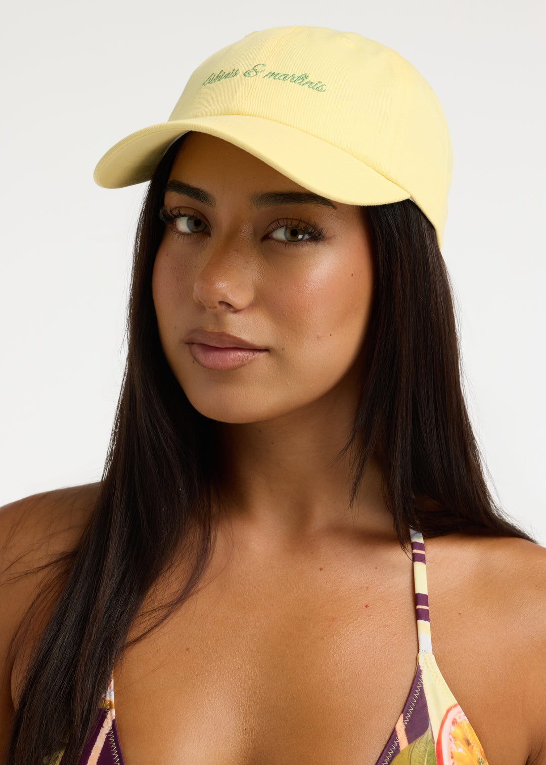 Model with long black hair wearing a light yellow Martini cap made from 100% cotton twill with ‘bikinis & martinis’ in silver cursive, paired with a Passiontini bikini top featuring a tropical passionfruit and floral print. Visible from head to chest.
