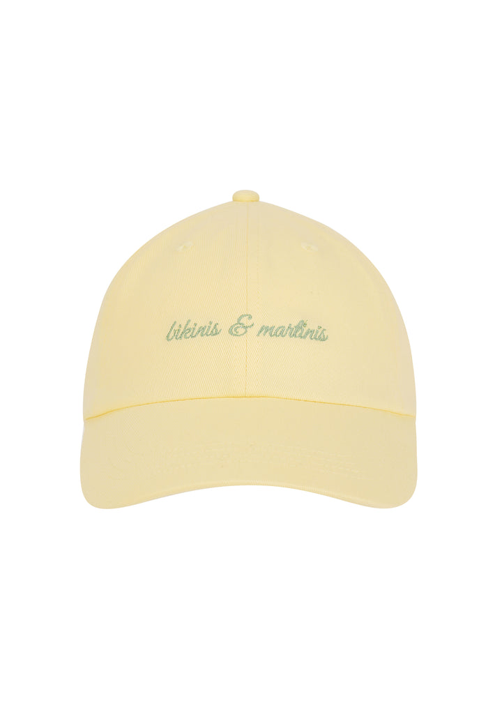 Light yellow Martini cap with the words "bikinis & martinis" in silver cursive on the front, made from 100% cotton twill, classic baseball cap design, against a white backdrop.